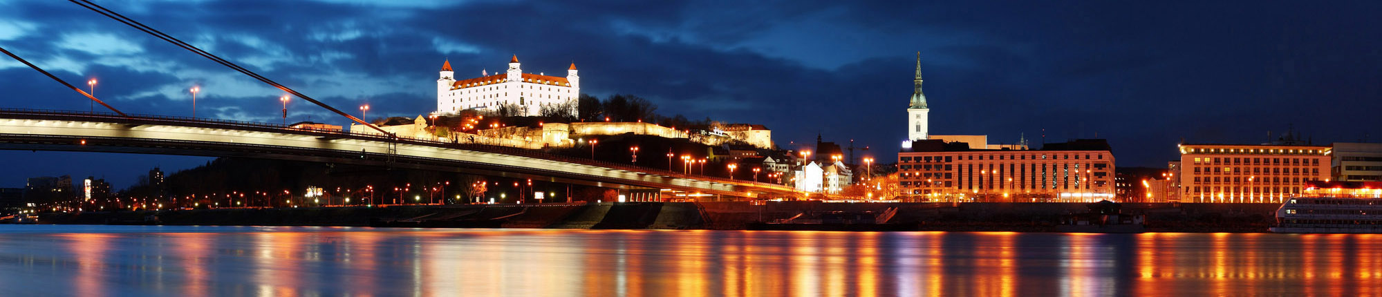Панорамний вид на Братиславу в сутінках, який включає міст, освітлені будівлі, включаючи замок на горі, та відображення на воді. 