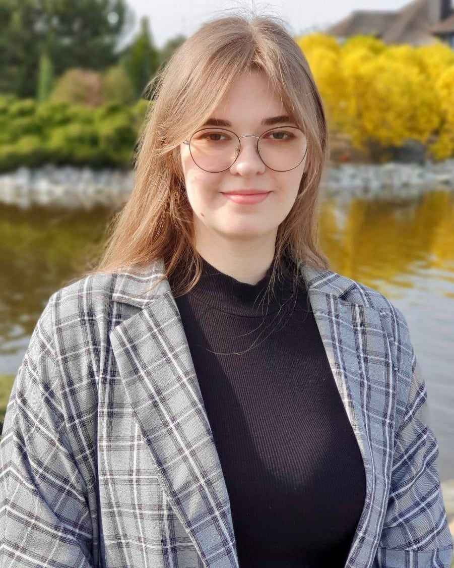 На фото зображена Єлизавета Турська – студентка Університету імені Альфреда Нобеля в окулярах на вулиці біля озера. Вона посміхається і одягнена в піджак 