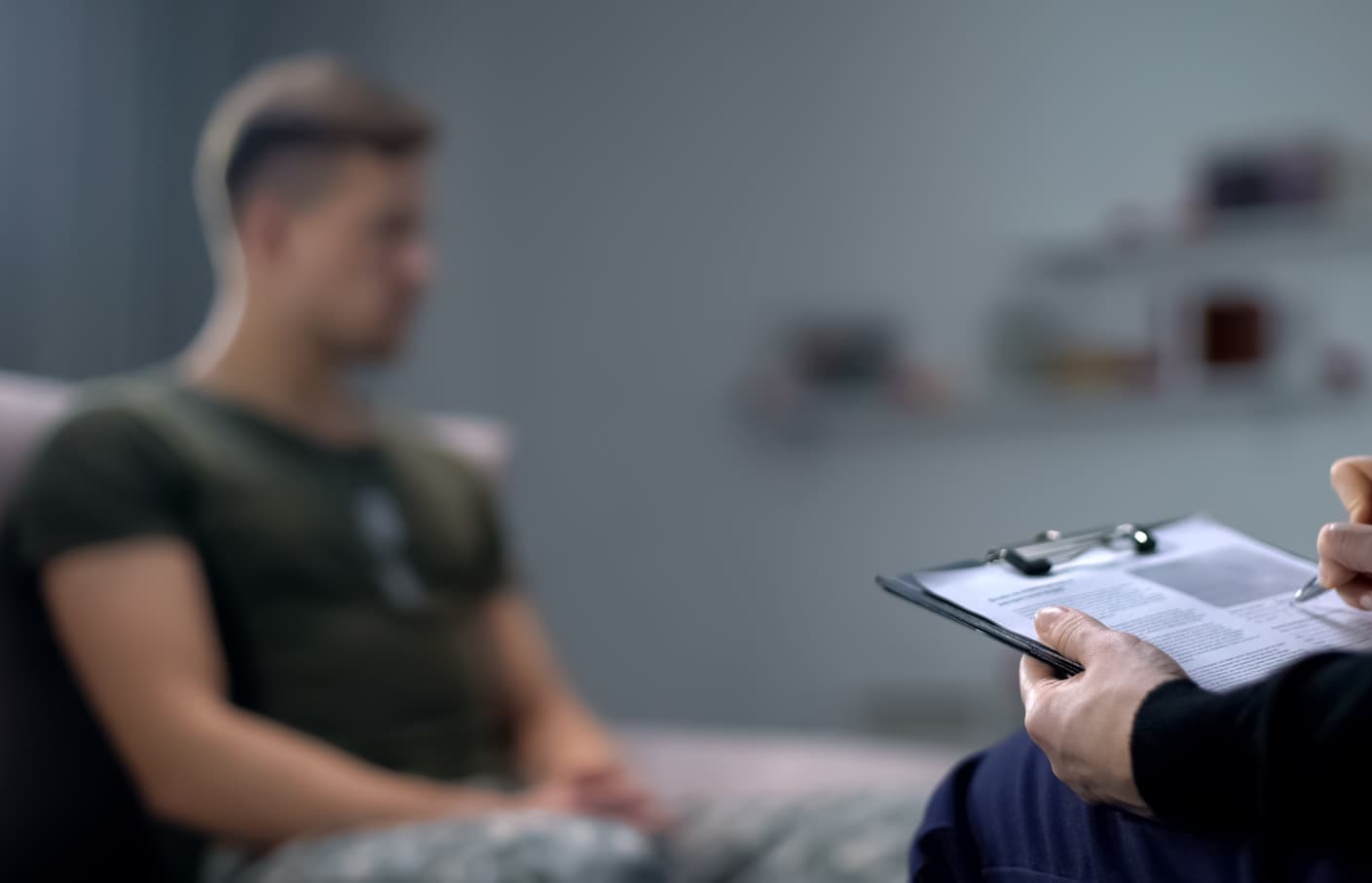 A blurred young man in a military uniform sits on a couch in a therapy session, while a therapist, whose hands are visible, takes notes on a clipboard. The setting appears to be a counseling or medical office with a neutral-toned background.