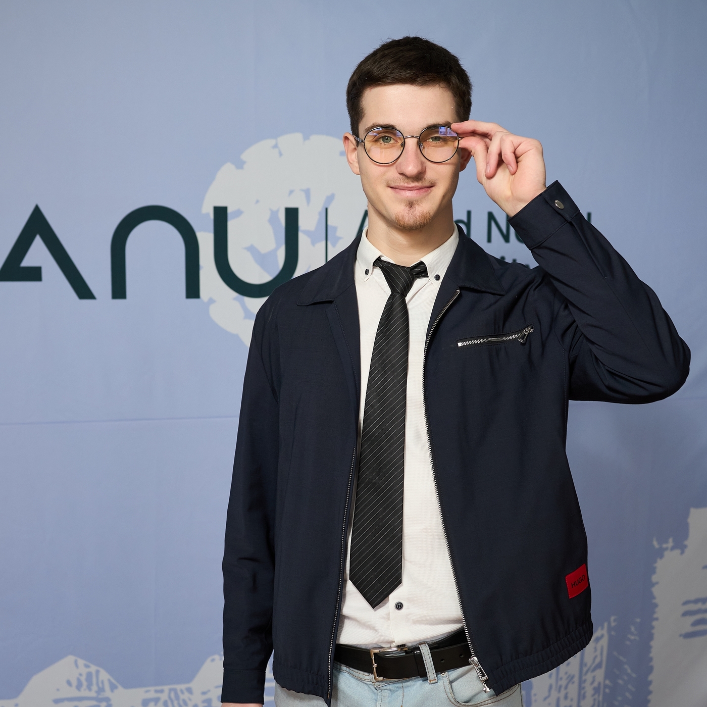 Молодий чоловік у діловому стилі з окулярами позує на фоні банера Університету імені Альфреда Нобеля, тримаючи рукою оправу окулярів
